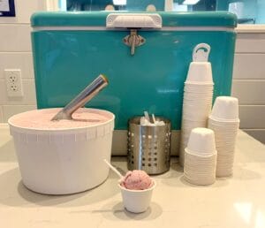 This is a picture of turquoise retro cooler with a half tub of strawberry ice cream and a scoop, cups, and spoons. 